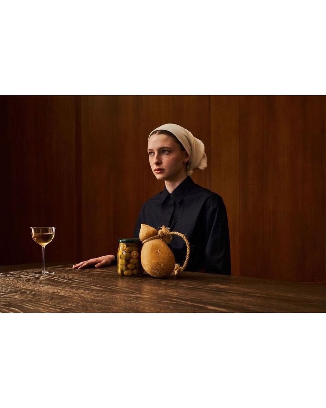 Young woman in headscarf at rustic table with wine glass, olives, and cheese. Soft indoor lighting, medium shot, studio backdrop.