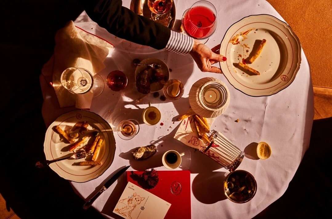 Overhead shot of a dining table with plates of food, glasses of drinks, silverware, and an adult human hand reaching for a plate.