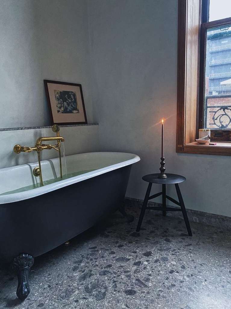 Freestanding black bathtub in a bathroom with dark gray terrazzo floor, natural light from a window with wooden trim, and wall art.