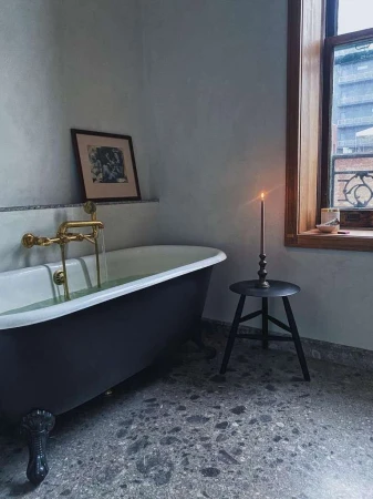 Freestanding black bathtub in a bathroom with dark gray terrazzo floor, natural light from a window with wooden trim, and wall art.