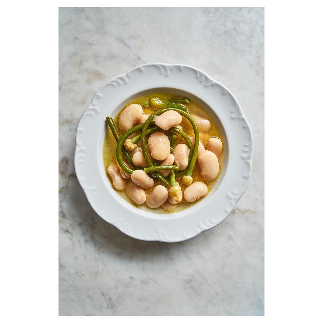 Overhead shot of white beans with green vegetables in broth, served on a white decorative plate, atop a marble surface.