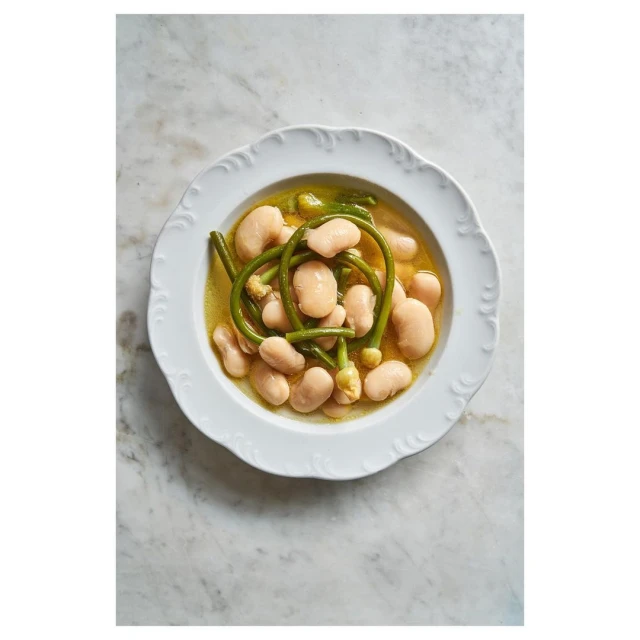 Overhead shot of white beans with green vegetables in broth, served on a white decorative plate, atop a marble surface.