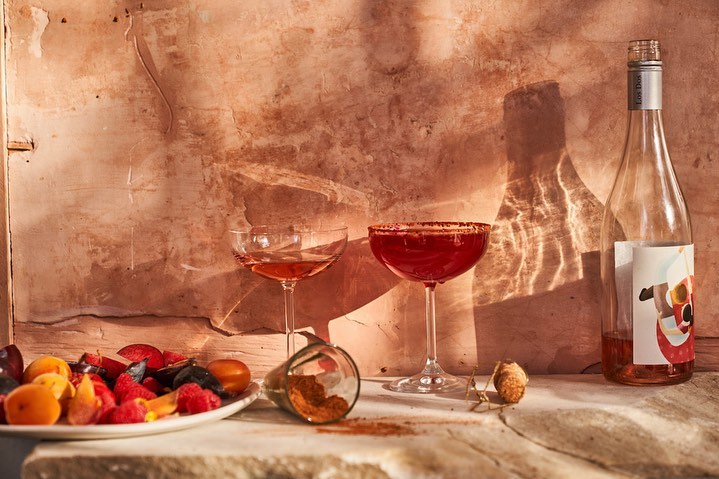 Still life shot of two coupe cocktail glasses, one with a red rim, alongside a bottle and a plate of mixed berries.