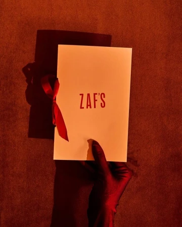 A gloved hand holding a white rectangular card with red text Zaf's and a red ribbon tied on the side