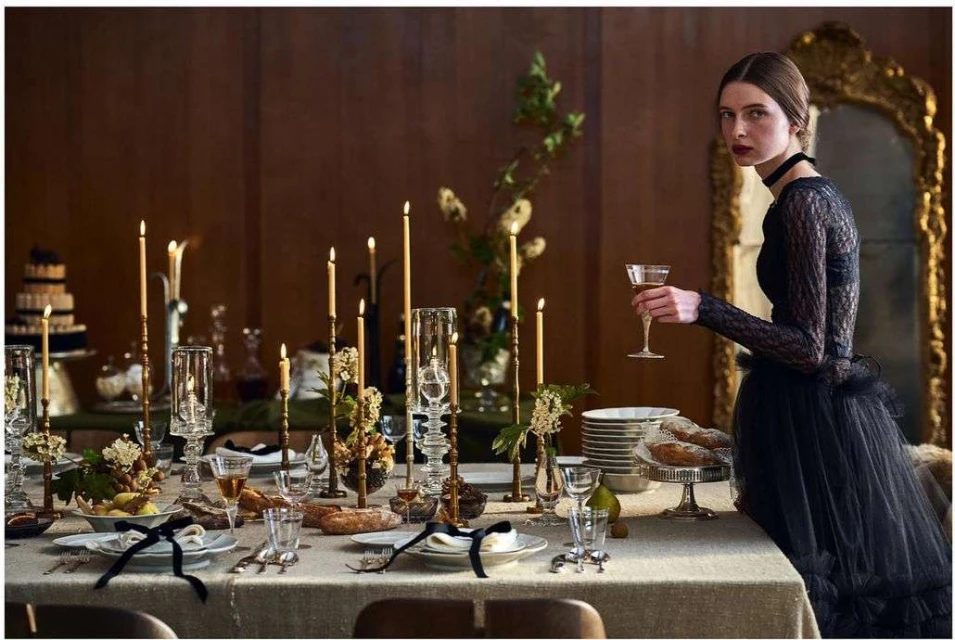 Woman in a black gown holding a cocktail glass at a candlelit table set for a formal dinner, with wood paneling in the background.