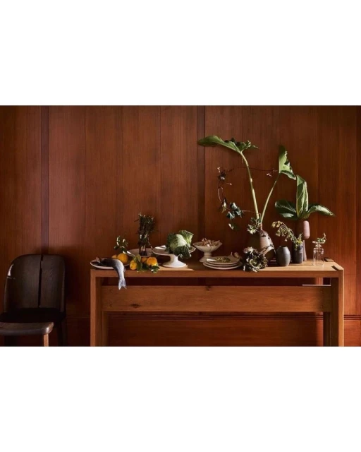 Still life composition features a wooden table adorned with fruit, tableware, and leafy plants, set against a paneled wood wall backdrop.