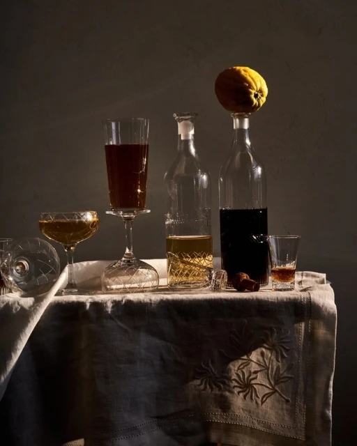 Elegant still life captures an array of glass liquor bottles and cocktail glasses filled with assorted alcoholic beverages on a tablecloth.