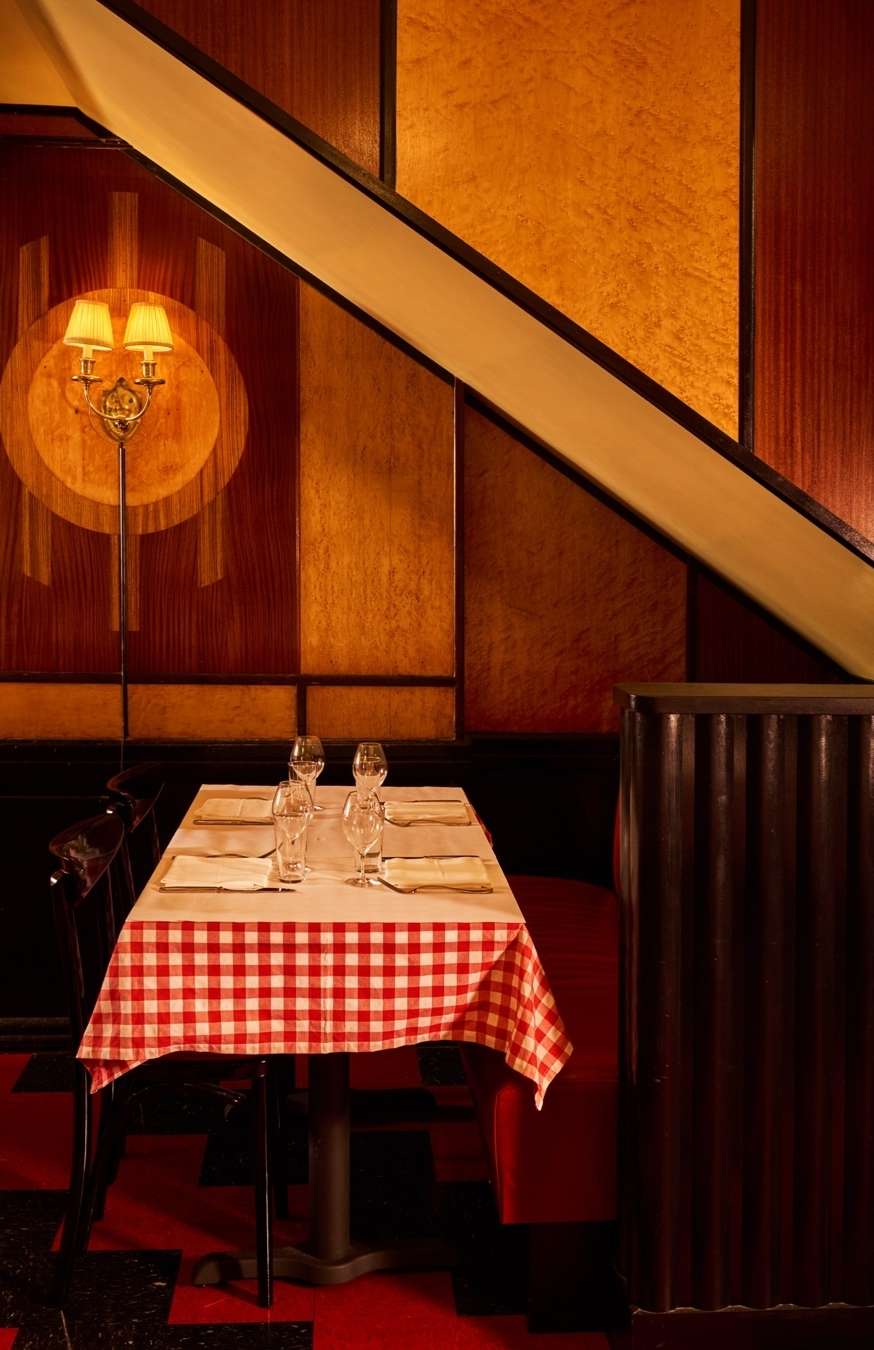 Restaurant interior featuring a set table with a checkered tablecloth, wine glasses, and patterned wood paneling under a staircase.