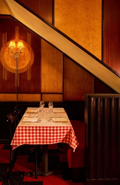 Restaurant interior featuring a set table with a checkered tablecloth, wine glasses, and patterned wood paneling under a staircase.
