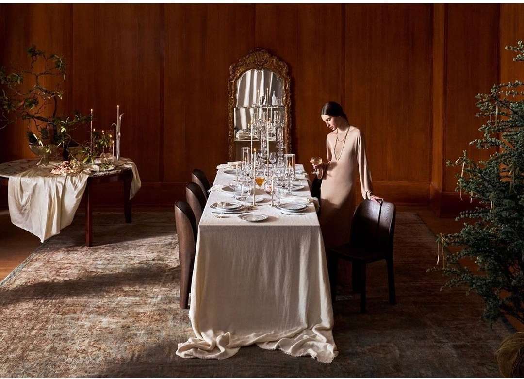 A woman stands by a long formal dining table set for a dinner party, featuring crystal glasses, candles, plates, and dark wood accents.