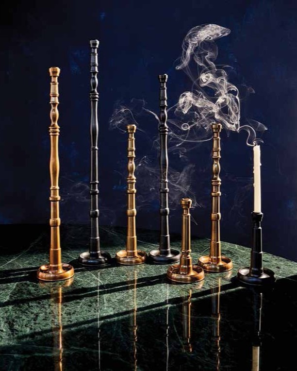 Still life featuring a collection of black and gold candlesticks of varying heights on a green marble surface with smoke and a candle.