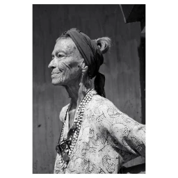 Monochrome portrait of an elderly woman wearing a patterned headscarf and layered beaded necklaces by photographer Gentl And Hyers