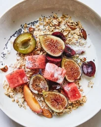 Bowl of breakfast cereal with milk, sliced fresh figs, plums, watermelon chunks, and almonds photographed from above.