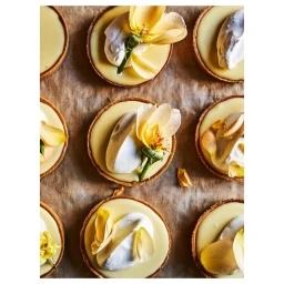 Top-down view of miniature lemon tarts topped with cream and yellow flower petals on parchment paper.