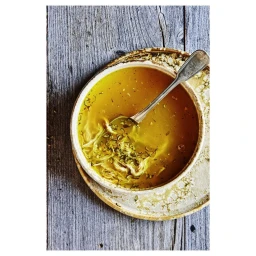 A ceramic bowl of golden broth with herbs and a spoon, resting on a wooden surface.