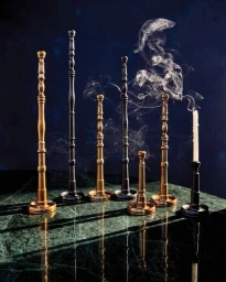 A collection of tall brass and black candlesticks on a dark green marble surface with rising smoke against a dark textured background.