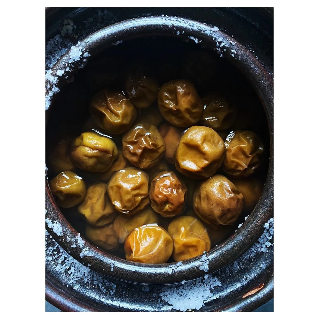 Round yellow salted plums curing in a dark ceramic jar covered with coarse salt crystals around the rim.
