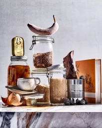 A still life arrangement of various food jars, a tin, a cup, blackberries, and an overripe banana balanced on top.