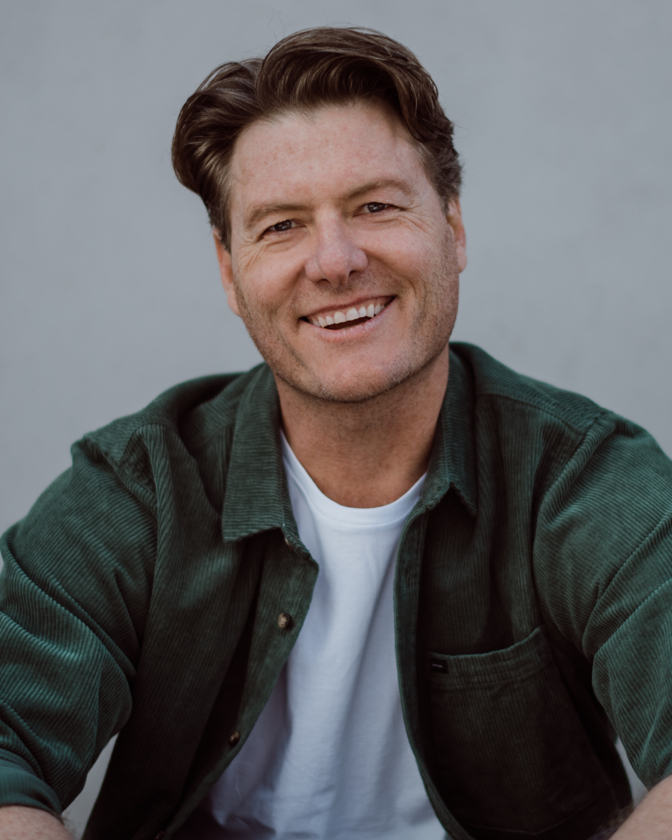 Close-up portrait of smiling adult male with brown hair, wearing a green corduroy shirt and white tee in front of a neutral background.