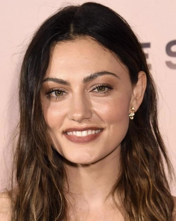 Close-up portrait of a smiling brunette woman with long wavy hair, wearing minimal makeup and gold earrings, against a muted pink backdrop.