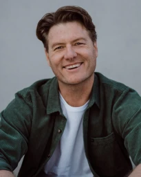 Portrait of a smiling man in his 40s wearing an unbuttoned green corduroy shirt over a white t-shirt against a plain grey background.