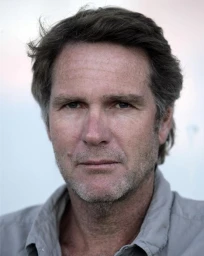 Close-up portrait of a middle-aged man with dark hair, light eyes, and light stubble wearing a grey collared shirt against a pale backdrop