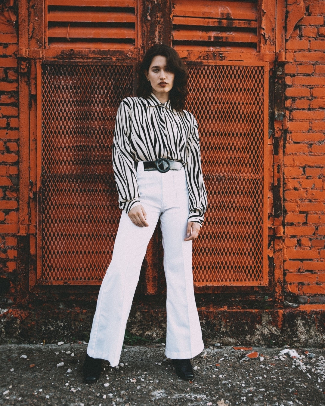 A young woman in a zebra-print blouse and white slacks stands in front of an orange metal door and brick wall, in a fashion portrait.