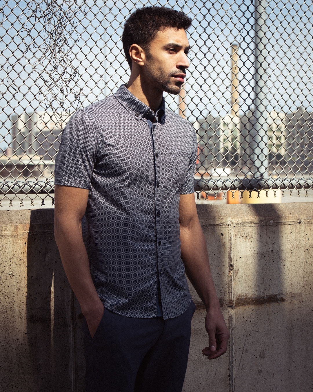 Adult male model wearing patterned button-down shirt poses against a chain-link fence with city buildings and a smokestack in background.