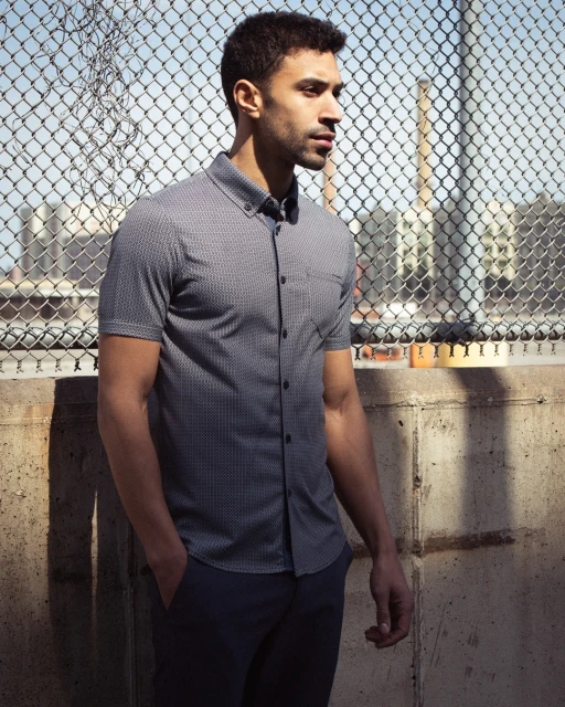 Adult male model wearing patterned button-down shirt poses against a chain-link fence with city buildings and a smokestack in background.