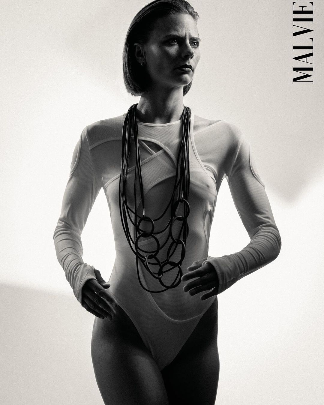Studio shot of an adult woman wearing a white bodysuit and black necklace, set against a plain background with the 'Malvie' logo.