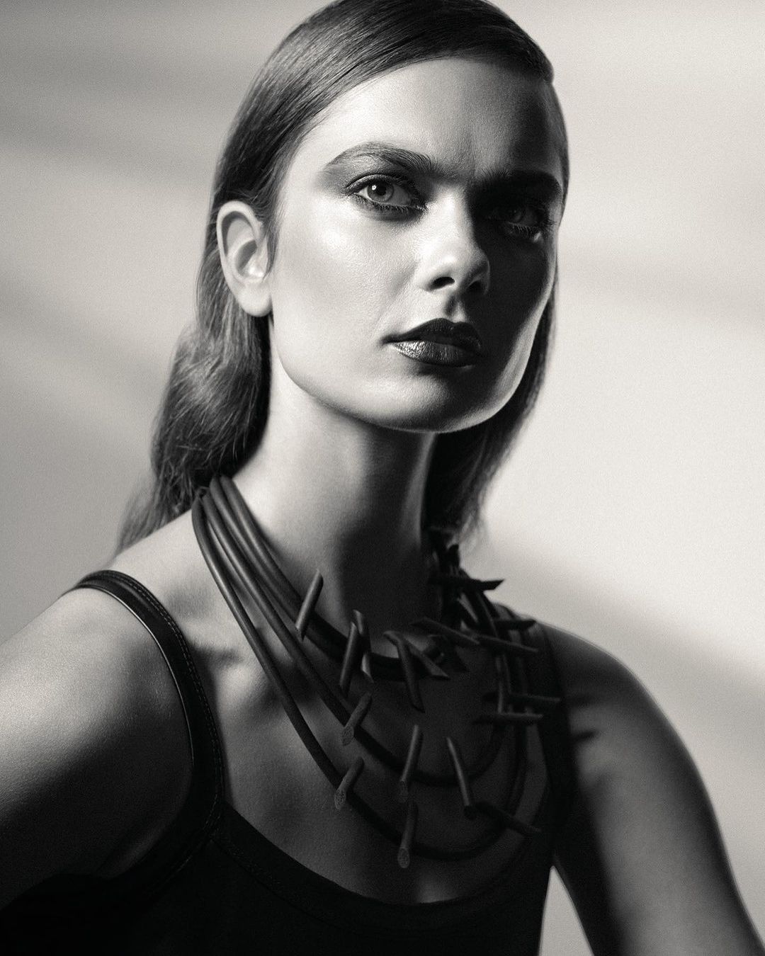 Monochrome portrait of an adult woman with slicked-back hair, wearing a contemporary necklace, and posing in a photo studio setting.