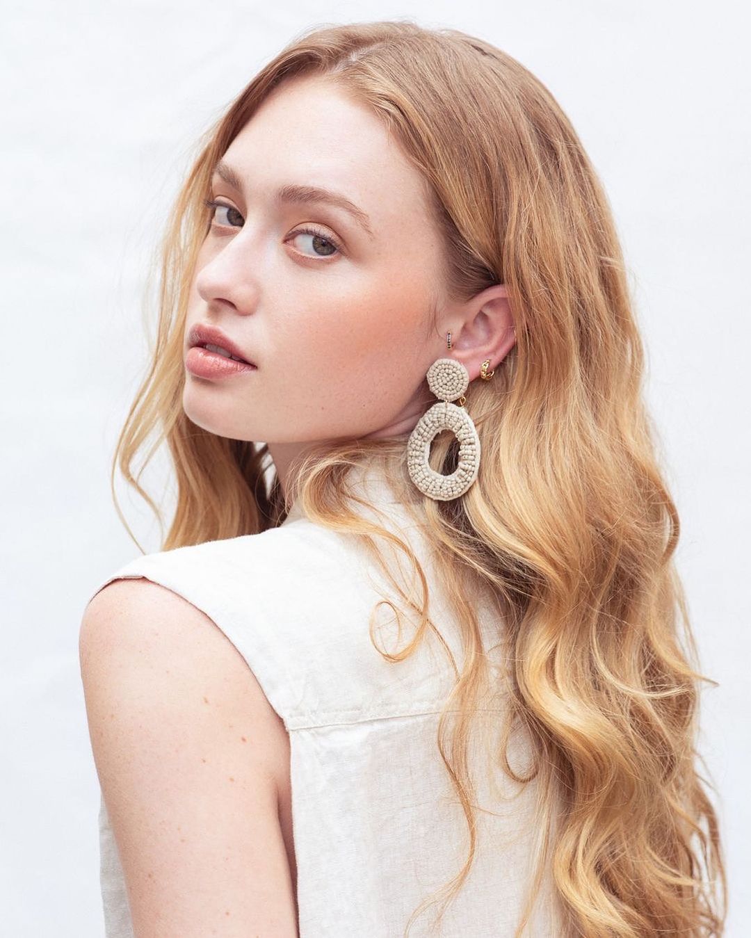 Close-up of an adult woman with long blonde hair wearing woven dangle earrings and a light sleeveless top against a white backdrop.