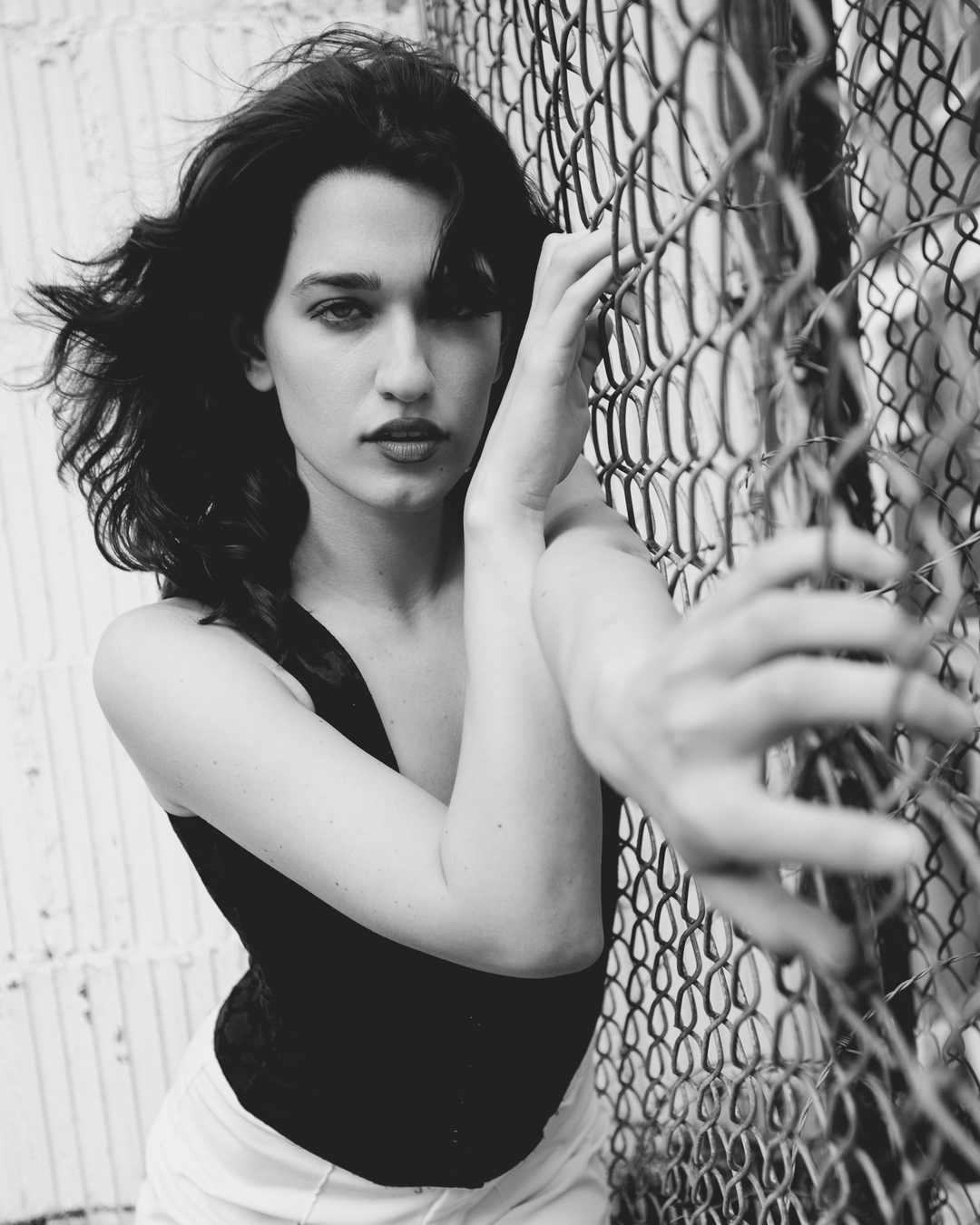 Black and white portrait of a woman posing near a chain-link fence, wearing a tank top and light-colored pants, with wind-blown hair.