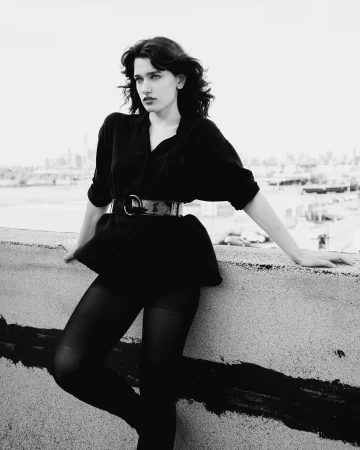 A woman in a dark shirt and belt leaning against a concrete ledge on a rooftop with a city skyline in the distance.