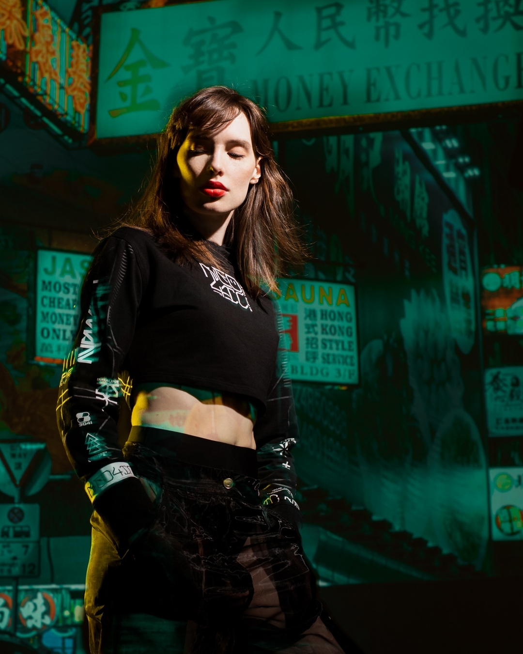 Stylish woman in a black crop top with sleeves poses in front of blurred signs at night with teal, yellow, and orange colors.