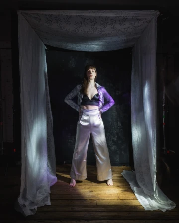 Full-length shot of a woman posing confidently in a silver jacket and pants under a draped white cloth, set against a dark background.
