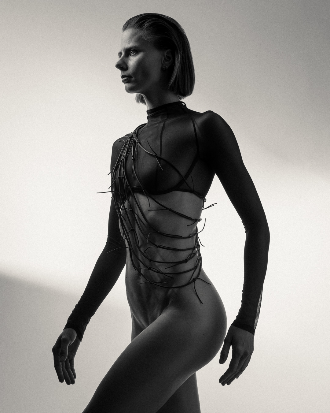 Monochrome portrait of a woman in sheer clothing with decorative elements, posing in a studio with a neutral background.