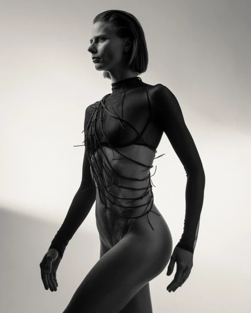 Monochrome portrait of a woman in sheer clothing with decorative elements, posing in a studio with a neutral background.