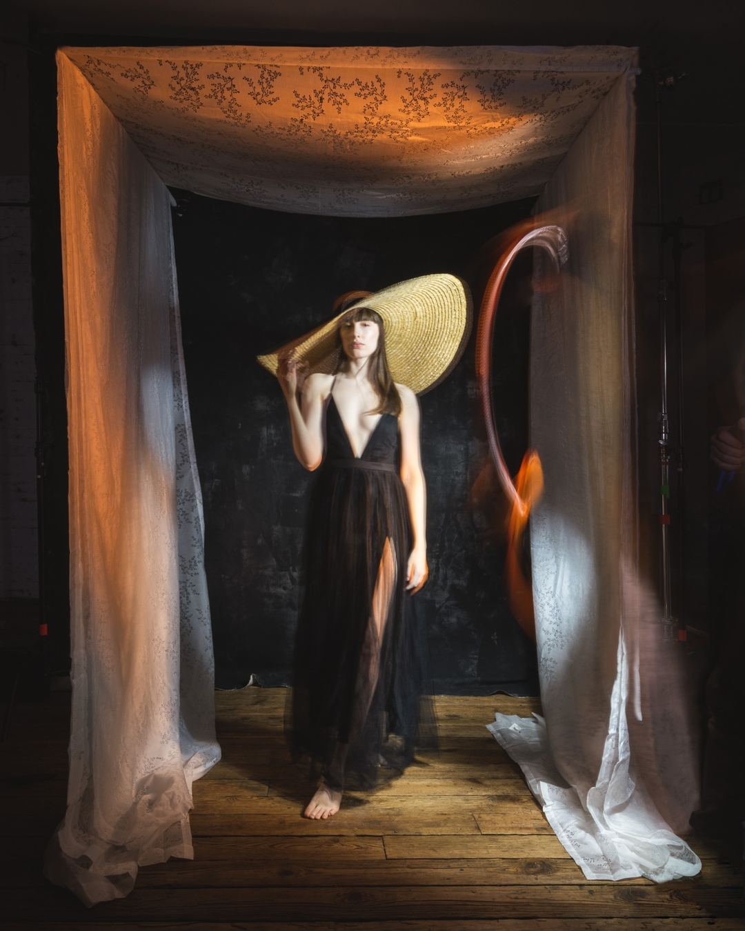 An adult woman wearing a black dress and an oversized sun hat standing barefoot on a wooden floor, in front of a dark backdrop.