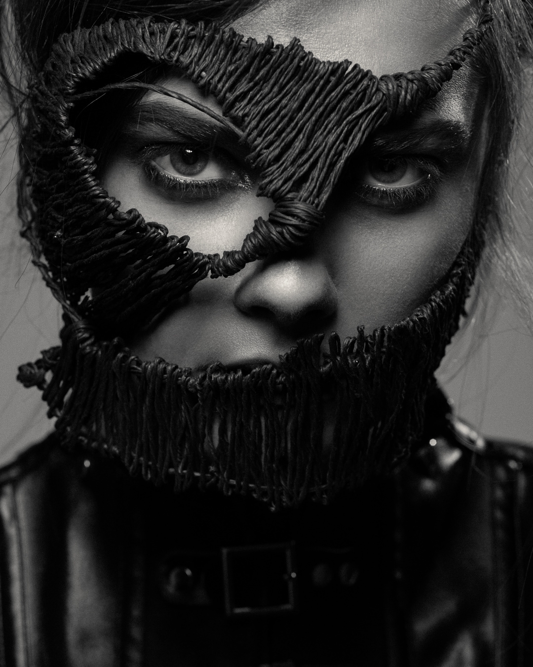 Striking black and white portrait of a woman with intricate thread mask, eyes focused, in a studio setting with soft lighting.