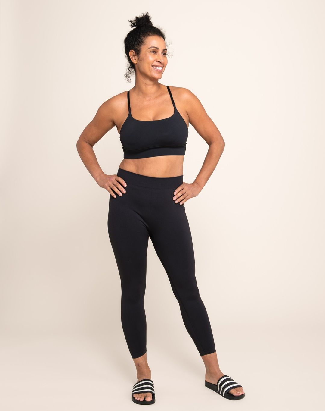 Full shot of an adult woman smiling in black athletic wear with her hands on her hips, posing in front of a neutral beige background.