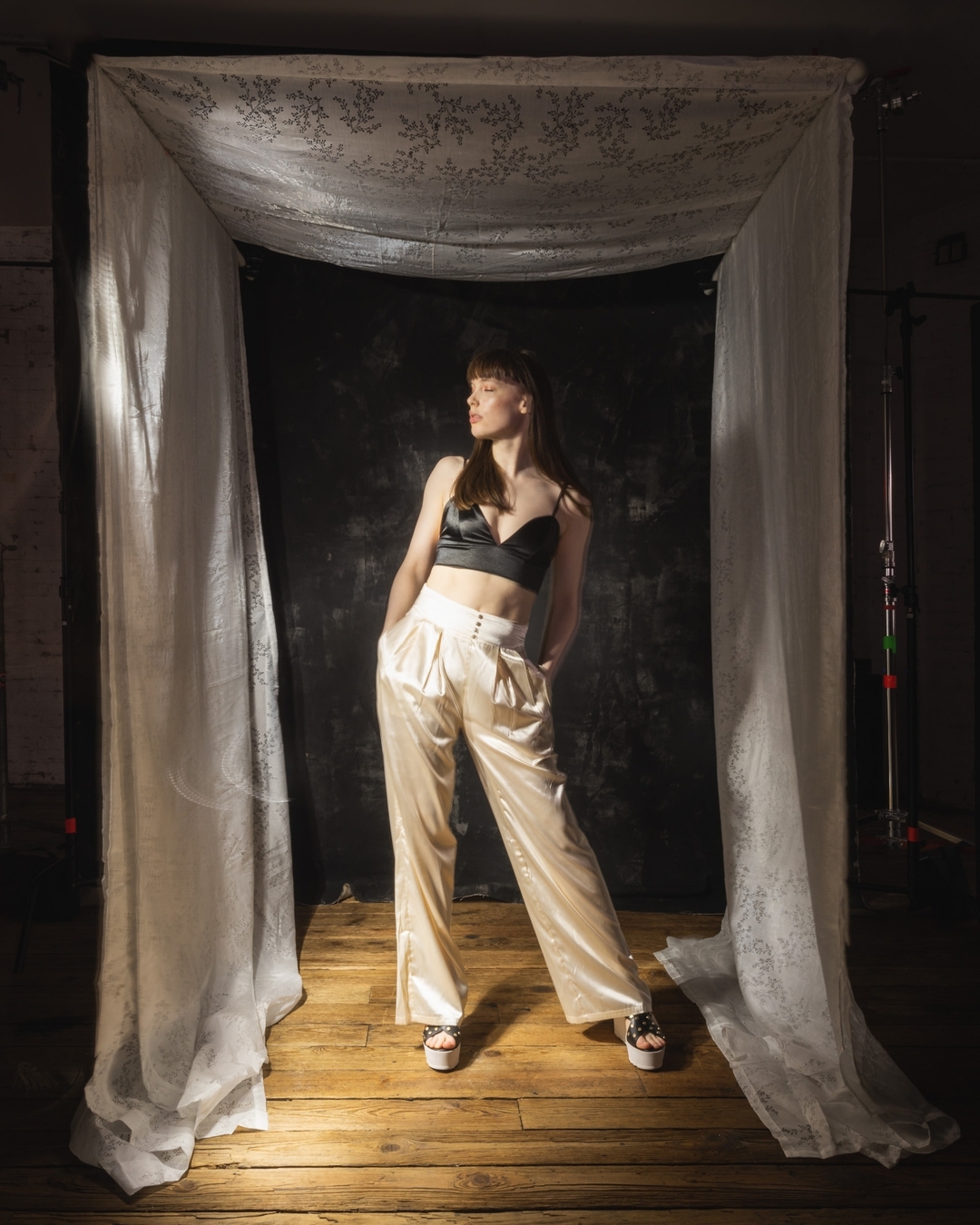 A stylish woman posing in a black crop top and cream-colored pants under a draped cloth in a studio setting, looking off to the side.