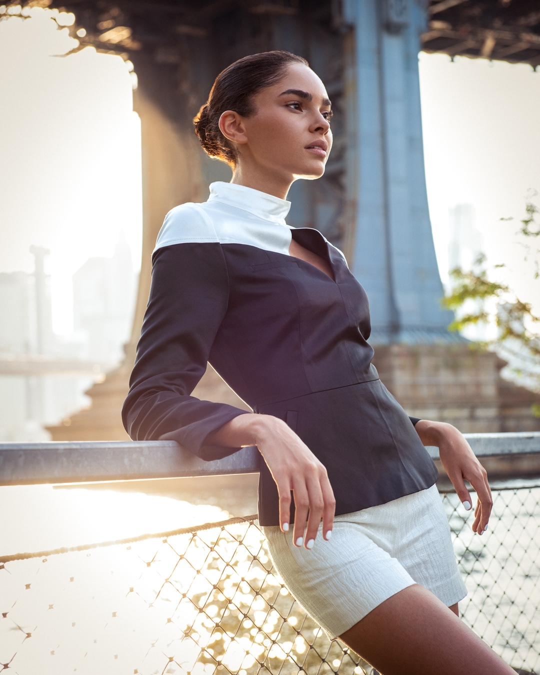 Woman in dark top with white collar leaning against a metal railing outdoors with bridge structure in background