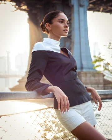 Woman in dark top with white collar leaning against a metal railing outdoors with bridge structure in background