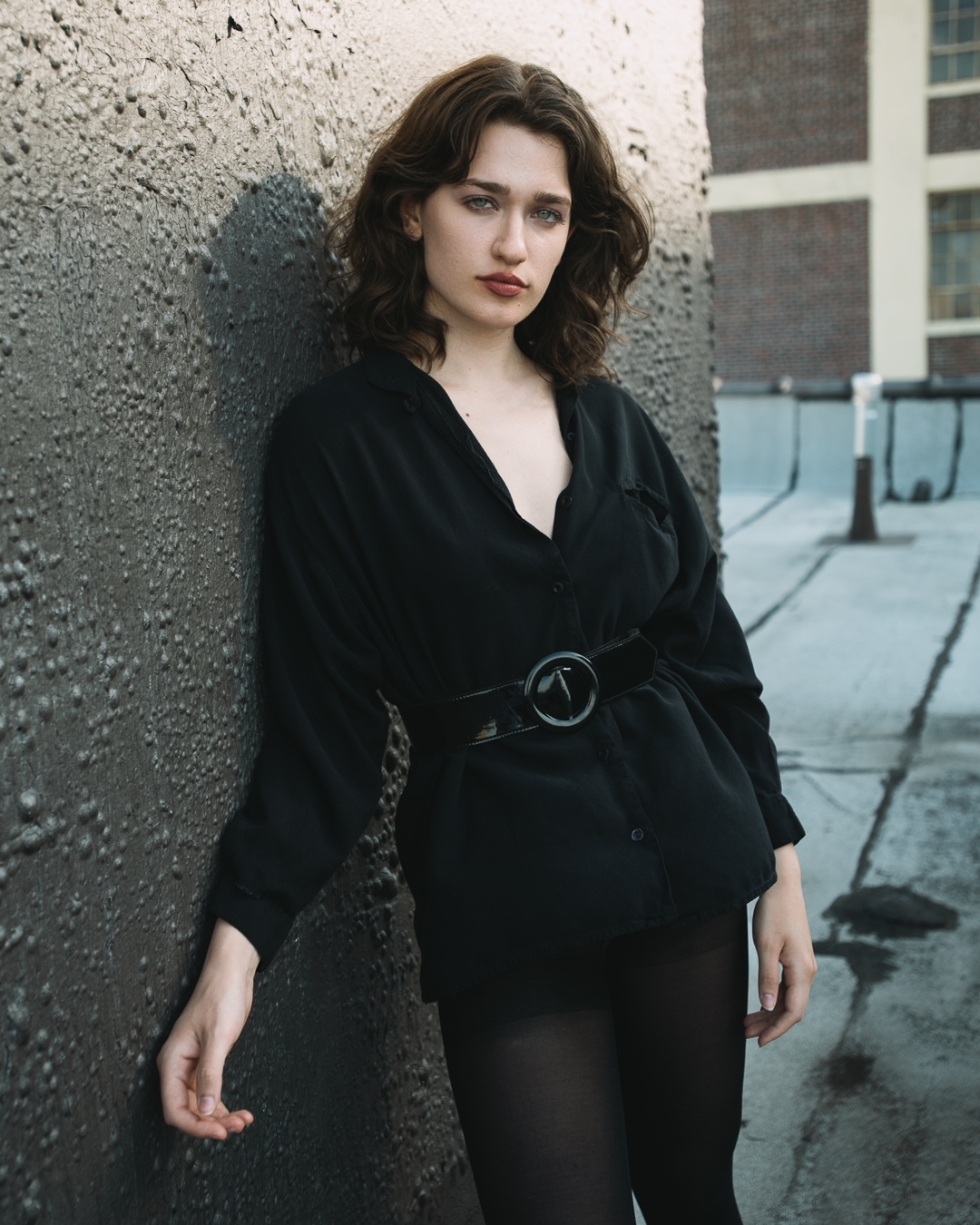 A woman poses against a textured gray wall outdoors wearing a black top, belt, and tights in a medium shot with natural lighting.