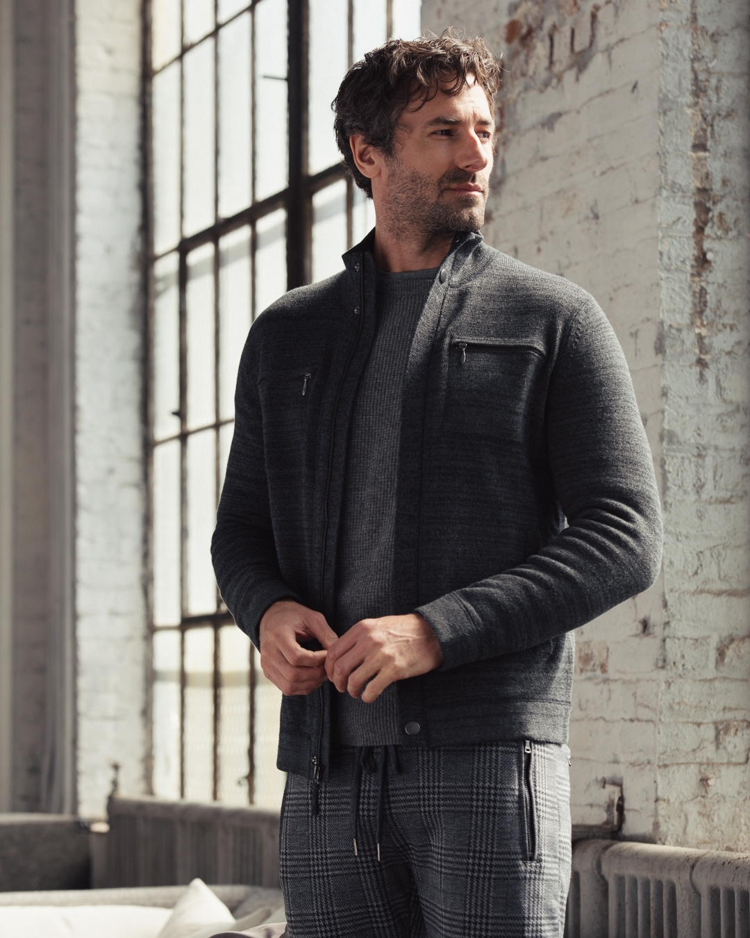 Adult male posing in gray jacket and plaid pants indoors near a window, showcasing contemporary men's fashion.
