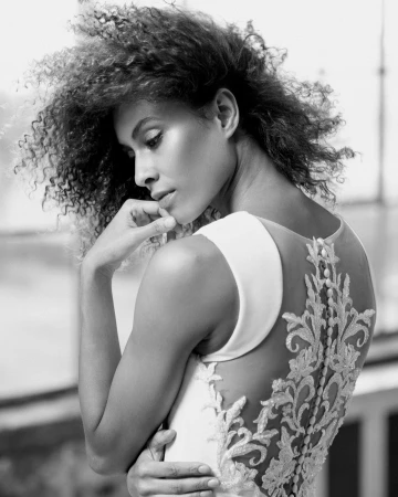 Black and white portrait of a woman with curly hair wearing a white sleeveless dress with detailed lace back embellishments.