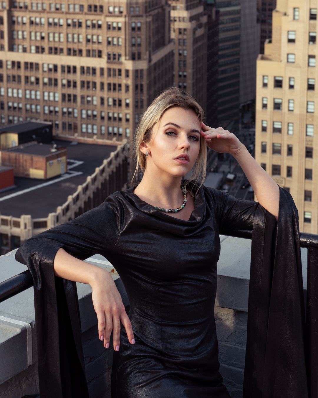 Blonde woman in long black dress poses on a rooftop with city buildings in the background in a medium shot, looking directly at the camera.