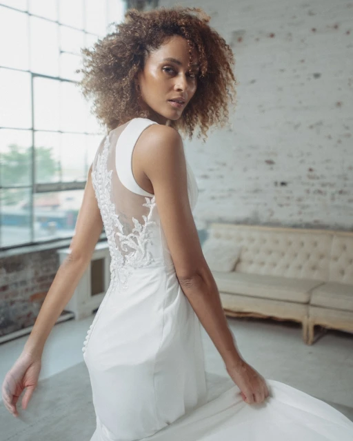Elegant African-American woman in a white wedding dress with a sheer back, posing in a bright studio with large windows and white brick.