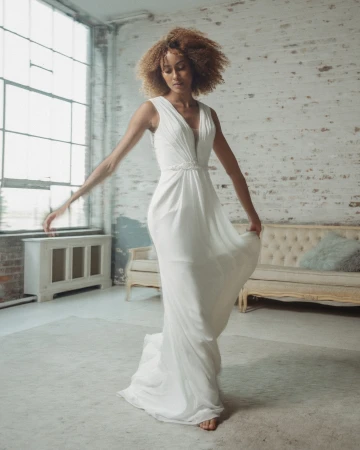 Elegant adult female in flowing white gown, barefoot in light-filled studio. Stylish bridal fashion, minimalistic aesthetic, soft natural lighting, and neutral palette.